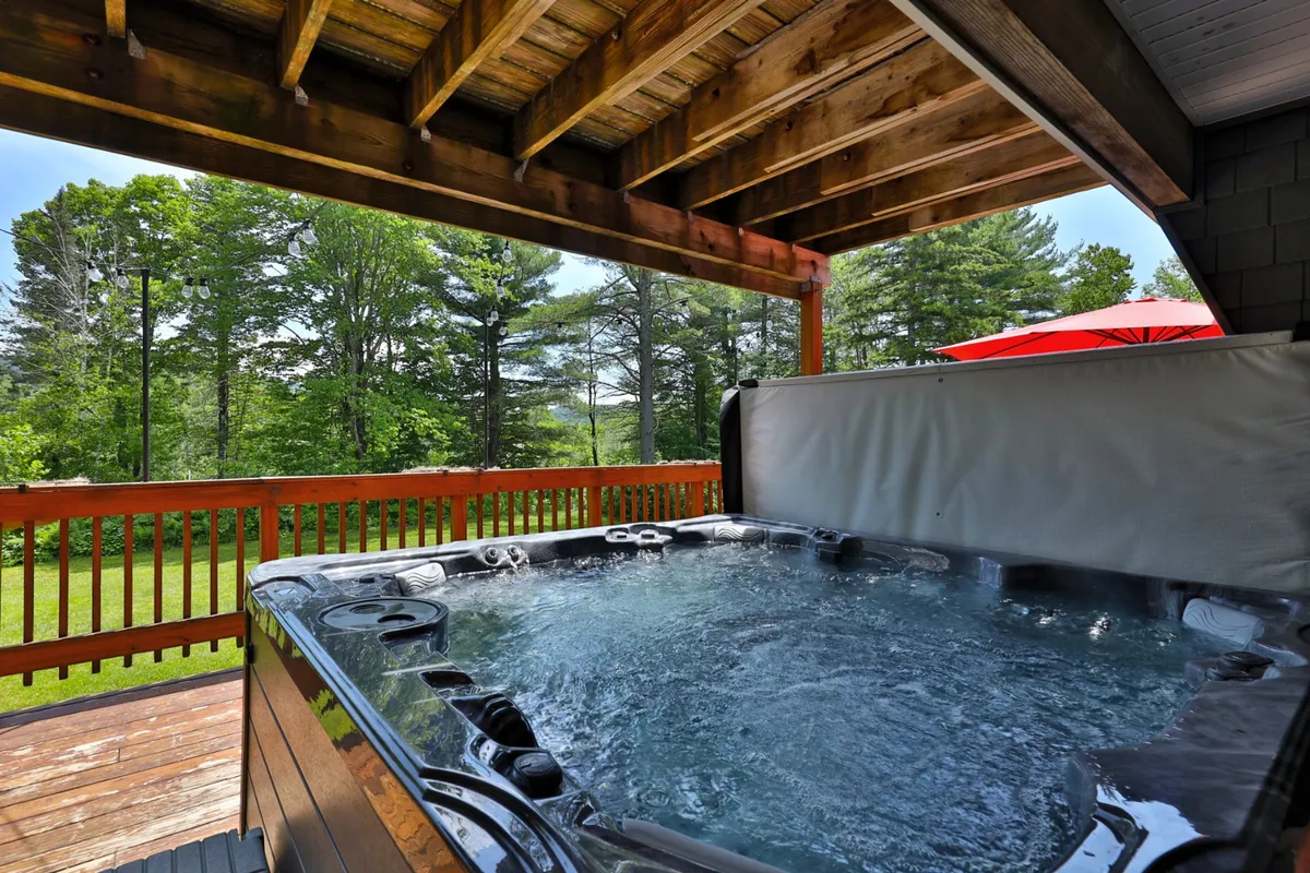 This image captures a cozy wooden deck nestled among mature forest trees, with a prominent hot tub and a red umbrella creating a focal point. The inviting outdoor area is set up for relaxation and socializing, with ample seating and tranquil nature views visible from the deck.
