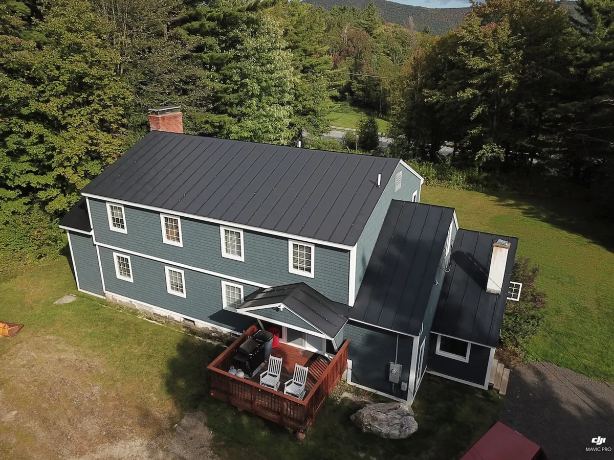 This exterior shot features the chalet's blue facade, a welcoming red deck, and mature trees encircling the property. The deck is set up for relaxation or socializing, with ample natural surroundings hinting at outdoor adventures nearby.