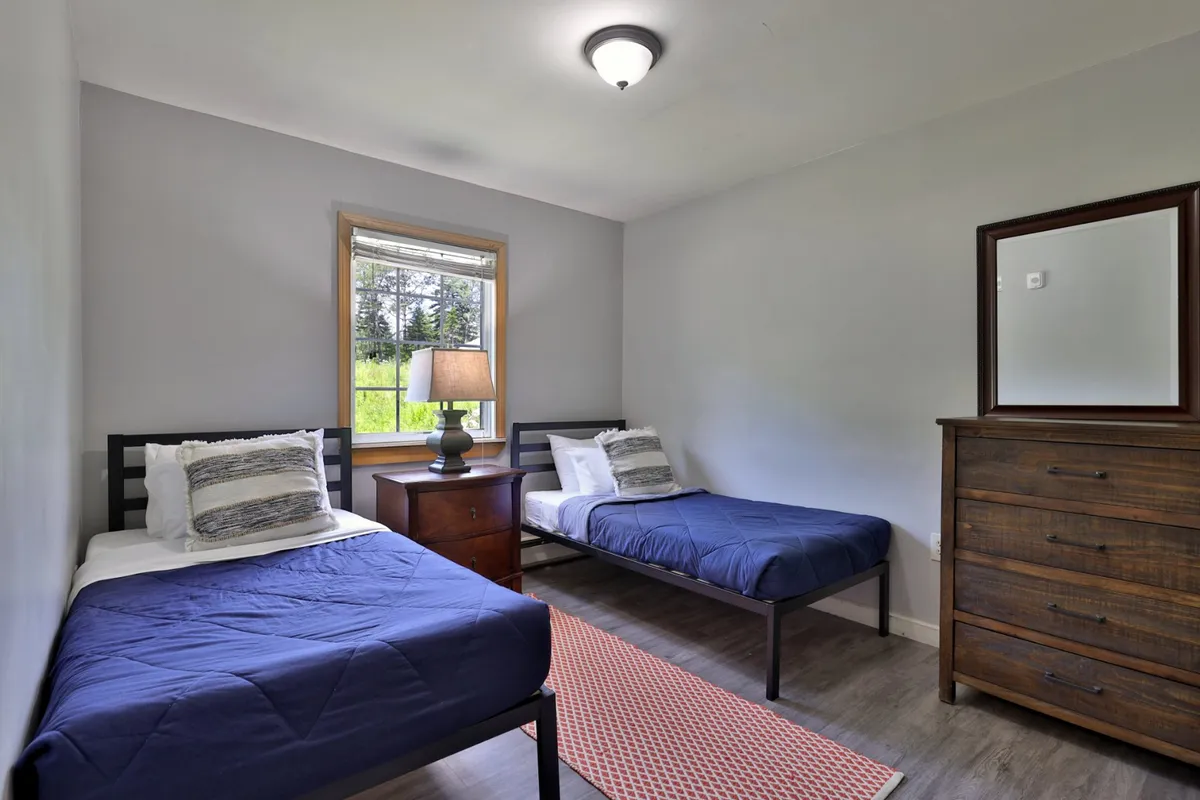 This image captures a cozy bedroom with two twin beds, a wooden dresser, a bedside lamp, and a window offering a peaceful view outside. The arrangement gives a sense of comfort and flexibility—perfect for children or guests to unwind after a day of adventure.