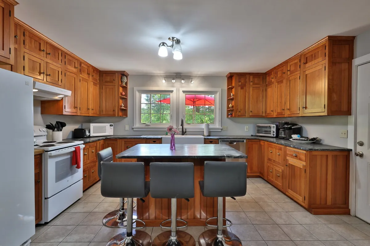 A bright, welcoming kitchen and dining area with a kitchen island, wooden cabinets, and stools. Windows let in natural light, with red umbrellas outside hinting at a connection between indoor and outdoor spaces. The setup is ideal for cooking, entertaining, and group meals.