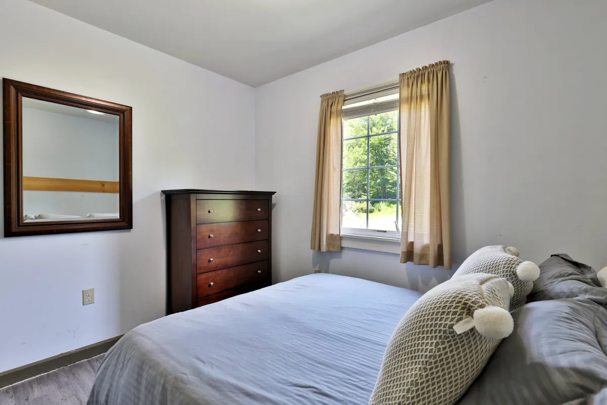 A sunlit bedroom featuring a soft bed, wooden dresser, mirror, and a large window. The setting is serene and inviting, ideal for parents or couples seeking a quiet, private retreat. Natural light enhances the sense of morning rejuvenation.