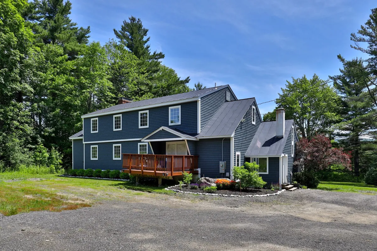 A serene exterior view of a two-story house with a large wooden deck, surrounded by greenery. The deck provides an inviting space for family gatherings, sunset viewing, or stargazing, blending relaxation with social possibility.